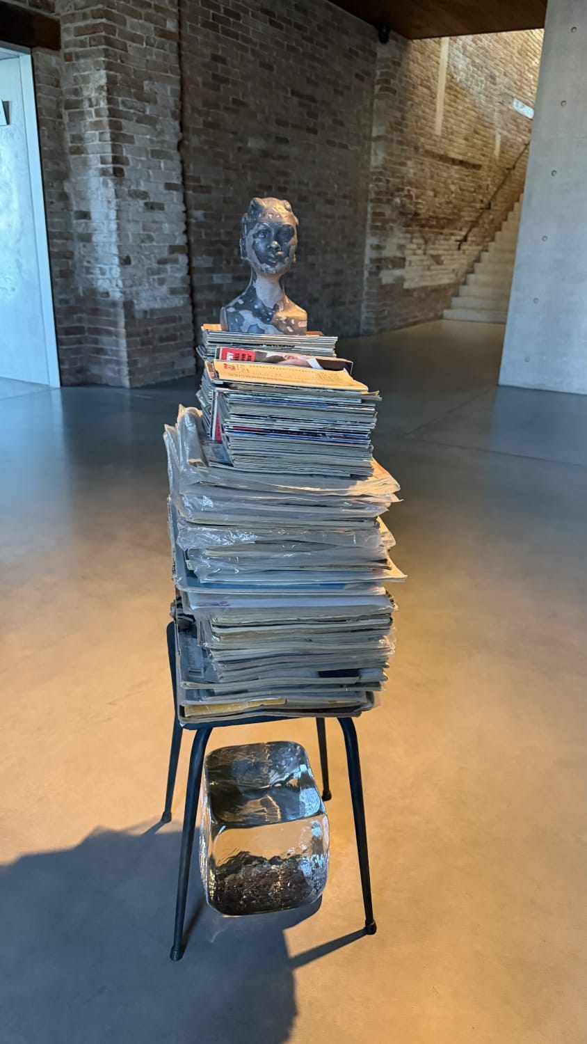 Instalação artística de Lorna Simpson na Fondazione Pinault, em Veneza, com pilha de revistas sobre cadeira, busto escultórico e elemento de vidro, explorando memória, arquivo e identidade na arte contemporânea
