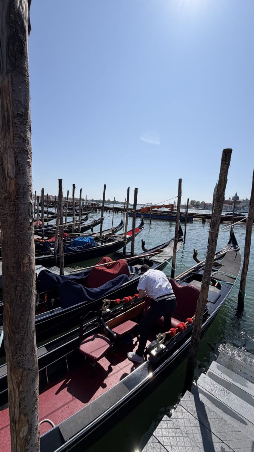 Gôndolas tradicionais ancoradas em canal de Veneza sob céu azul, com gondoleiro preparando embarcação, e vista para igrejas históricas ao fundo