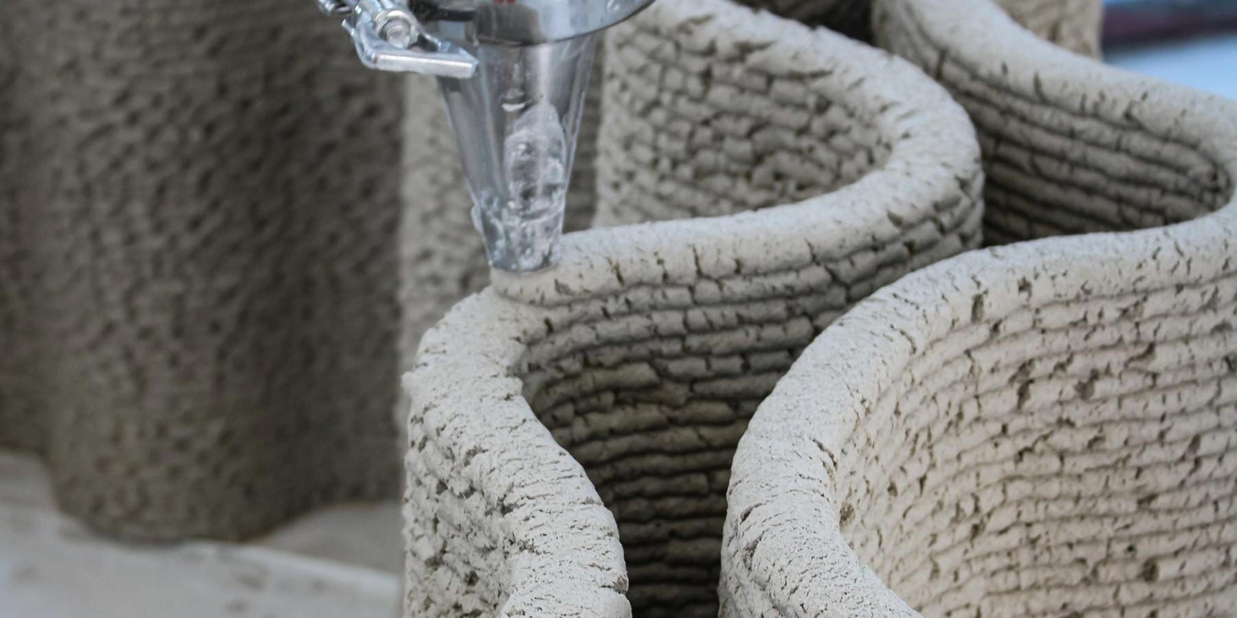 Bico de impressão 3D depositando o Corncretl em camadas sinuosas, formando parede em tom de areia