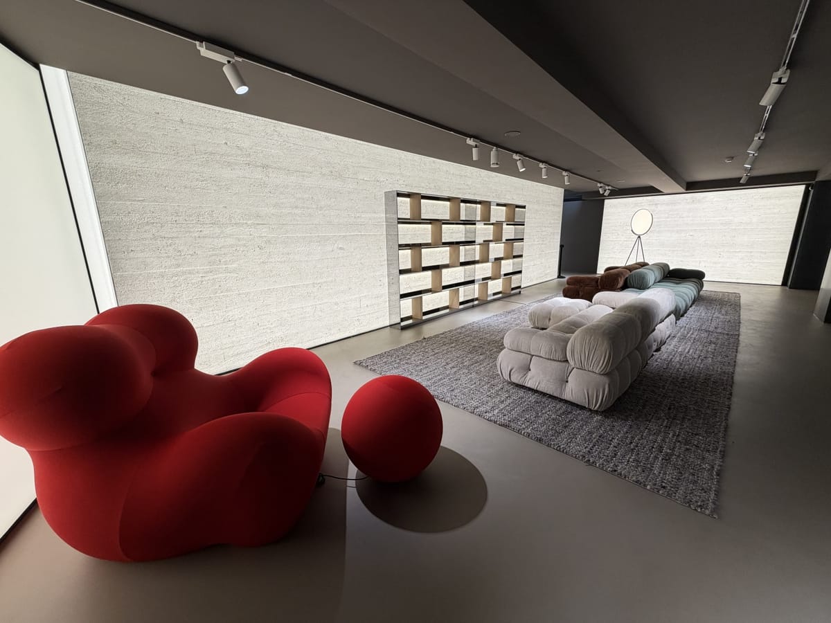 Estande contemporâneo na Semana de Design de Milão 2026, com poltrona vermelha escultural, sofás modulares em tons neutros, estante minimalista e paredes texturizadas 
