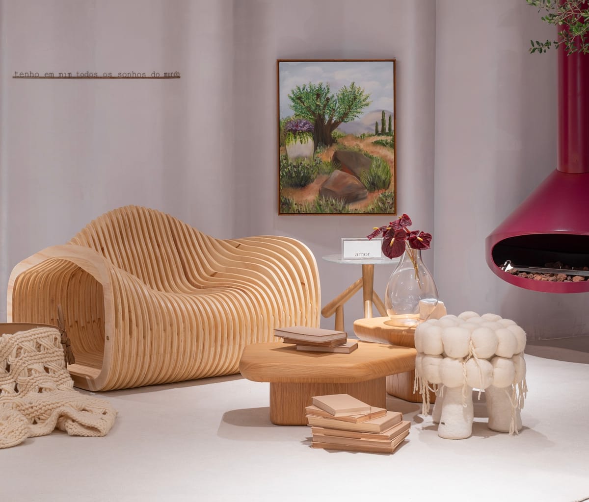 Ambiente da CASACOR BC 2026 com poltrona de madeira escultural, mesa orgânica, banco em lã e lareira suspensa rosa em decoração contemporânea acolhedora