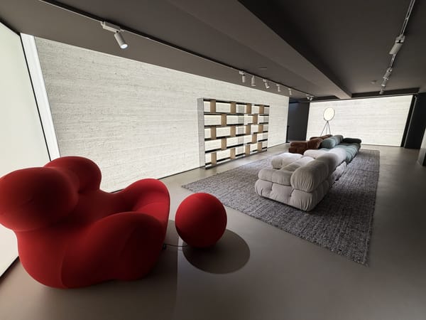 Estande contemporâneo na Semana de Design de Milão 2026, com poltrona vermelha escultural, sofás modulares em tons neutros, estante minimalista e paredes texturizadas 